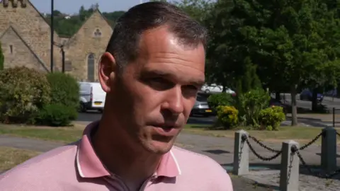Michael Harnett talks to a camera. He is clean shaven with short dark hair and is wearing a pink shirt