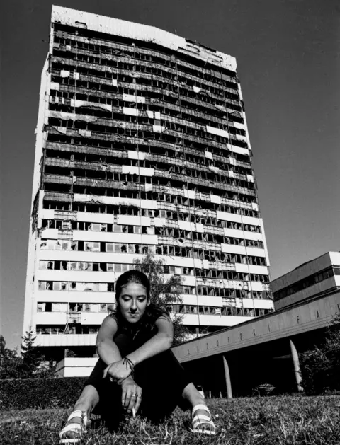 Chris Leslie A young woman sits on the ground with a war-torn tall building behind her