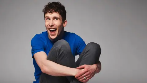 Matt Lockett Larry Dean, in a blue t-shirt and black jeans, smiling and looking out of the corner of his eyes with his hands wrapped around his knees 