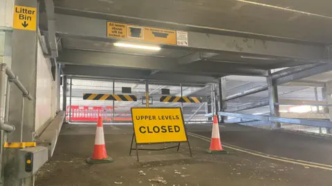 A car park interior. There are some gates that are closed at the top of the ramp, and a sign in the foreground that says "upper levels closed" next to some cones