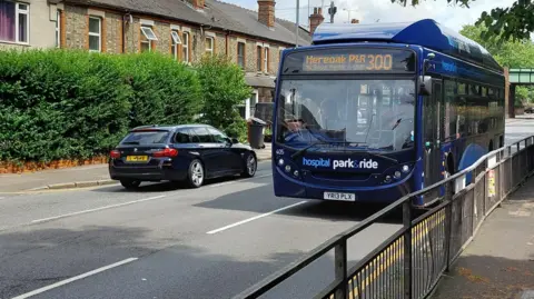 Reading Borough Council A bus along London Road, Reading, where a new bus lane will be established. It is a blue Hospital Park and Rode bus in a residential area. A dark coloured car is passing by it, travelling in the opposite direction. It is a sunny day.