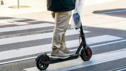 A person wearing trainers and trousers riding a black e-scooter across a pedestrian crossing 