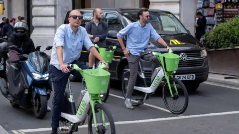 A black van, a motorcycle and two males on Lime bikes waiting behind a white line on a road, presumably for traffic lights to go green.