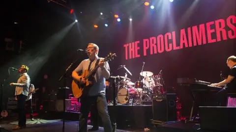 Getty Images The Proclaimers performing on stage. The two brothers are singing into mics and there are people playing the drums and keyboards behind them. There is a large projection saying The Proclaimers in red