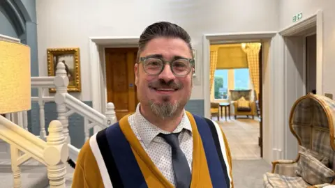 A man standing in a spacious room with a chair to his right and staircase to his left. He's wearing a white patterned shirt with a dark grey tie and a sweater over the top of it. He's got a big smile and is wearing prescription glasses.