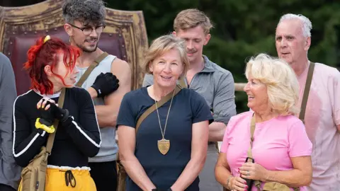 BBC/Studio Lambert/Euan Cherry Amanda Collier (centre) with fellow contestants looking at her during a mission on the Traitors show. She is wearing a navy t-shirt and a shield around her neck. She is smiling.