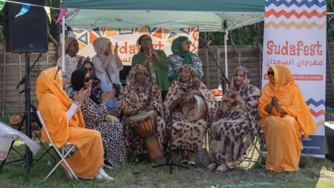 Sudafest CIC A group of Sudanese women are sat underneath a gazebo, outside in the sun. They are playing drums and singing together. Some are standing and some are sat down.