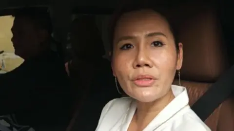 A woman wearing a white car sat in the passengers seat of a car. She is looking towards the camera out the window.