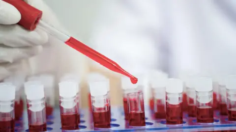 A close-up view of a gloved hand holding a dropper filled with red liquid above several laboratory vials, with a blurred clinical setting in the background.
