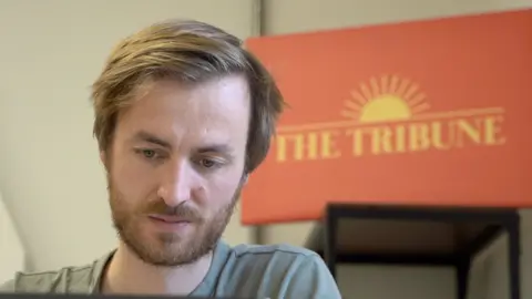 A man with blonde hair sits on a chair, with a red and white logo behind him