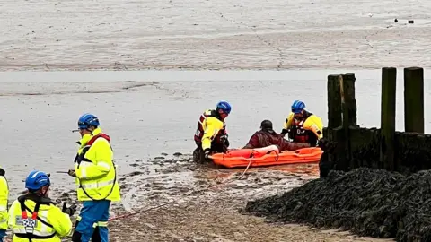 The man stuck in the mud with his back to the camera is helped by two rescuers in high visibility gear, life jackets and blue helmets other rescuers can be seen in the foreground