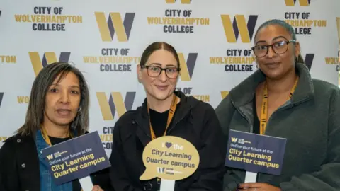 City of Wolverhampton Council Students and staff stand in front of a large poster advertising the City Learning Quarter as they hold small placards saying the same thing. The three women are smiling at the camera.