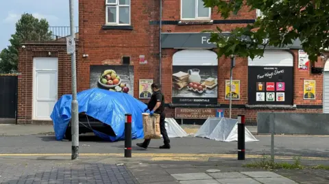 A police man carrying a large brown paper bag as he walks passed a car covered in a blue plastic sheet and two small silver tents