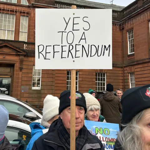 A man in a woolly hat stands holding a sign saying "Yes to a referendum"