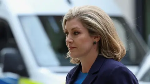 Penny Mordaunt is pictured walking outdoors. She wears a dark suit. 