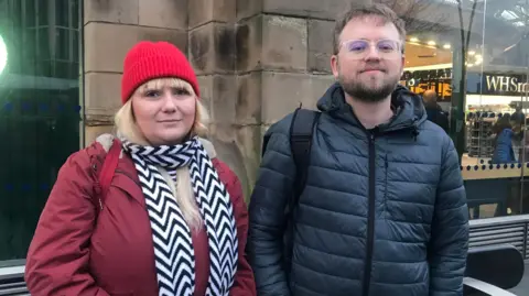 BBC/Victoria Scheer The pair stand in front of Sheffield train station, wrapped in winter coats. Ms Fitt-Stirling is wearing a black and white scarf and red hat, while Mr Partridge is wearing glasses.