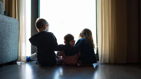 Backs of three small children sitting by window. The two bigger children are on the sides and touching the smaller child in the middle. There is a sofa on the side.