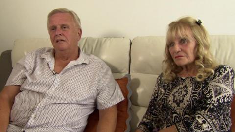 Chris and Karen Fleming sat on a cream leather sofa. Mr Fleming, on the left, has a white open collar shirt with circles on and has grey hair, while Mrs Fleming has shoulder-length blonde hair in curls with a dark pattern dress
