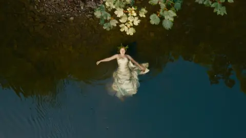 Megan Ruth-Trump Megan Ruth-Trump is floating in the river. She is seen from above. She is wearing a dress.