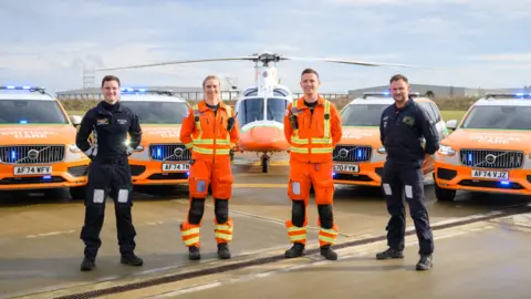 Team of Magpas air ambulance crew- three men and a woman- with two of them wearing orange uniform and the other two wearing black uniform, standing in a semi-circle in front of orange emergency vehicles and a helicopter. 
