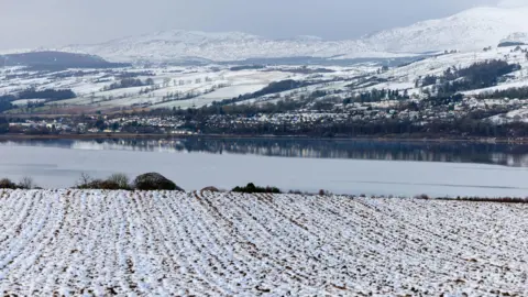 PA Media A snowy winter's day in Dingwall in the Highlands. Snow is covering all the grass, with a river behind and then hills beyond that, also covered in snow.