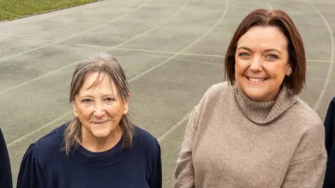 Two women are standing on a grey athletics running track with six lanes. On the left the woman is shorter with shoulder length grey brown hair and is wearing a navy blue sweater. On the right the woman is taller with short brown hair and a beige jumper