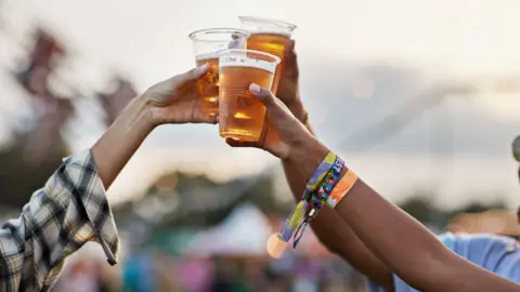 Three people cheers pints of beer in plastic cups, one has a rolled up sleeve and another has wristbands.