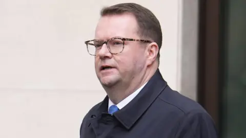 Conor McGinn, 41, who has short brown hair, glasses with tortoise-shell frames and is wearing a dark blue mackintosh coat, walking with a serious expression. 