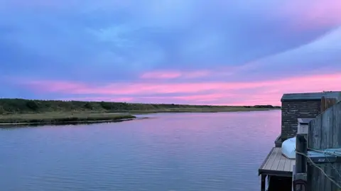 A sun sets over a wide body of water. It is lined with bushes. 