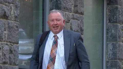 BBC Deputy Liam McKenna - A man with a grey suit on and a white shirt and a tartan tie. 