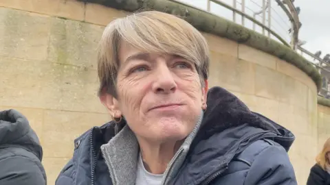 Robby West/BBC Trish Goodall is in the middle of the frame. She has short blonde hair and is wearing a grey T-shirt and jacket, and a blue coat. 