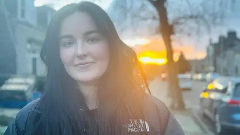 BBC Kate Finlay, young woman smiling at camera in street, with sun shining in the background.