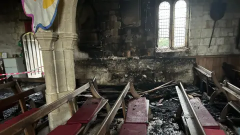 Pews inside a church with black fire marks on the wall. There are red cushions on the pews and a banner hangs from a pillar on the left.