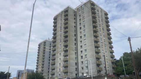 Two tower blocks. They are residential and have balconies. The render is cream but is visibly worn. The sky behind is grey.