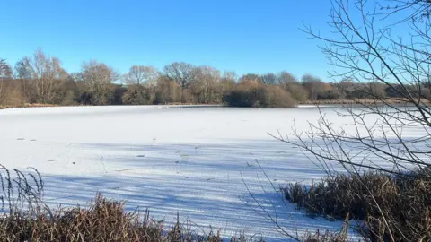 Frankie/BBC Weather Watchers A view of Orton Waterville in Peterborough on a sunny day. The lake is largely frozen over during cold weather. Trees and bushes surround the edge of the lake.