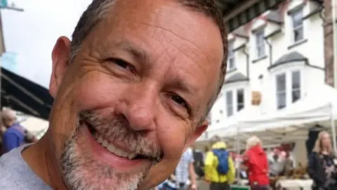 Terry Blake Michael Blake smiling while sitting outside at what appears to be a market. He has a grey goatee and short hair.