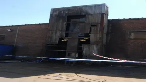 HSE A dark brick building with a corrugated steel tower attached to the side. Blue-and-white-striped tape and red-and-white-striped tape are in front of the building.