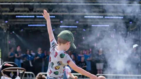 Nene Park Trust A child wearing a grey T-shirt with superheroes on it and a green cap dancing with his hands in the air. He is facing a stage with performers on and dry ice smoke.