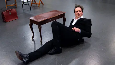 Myles Morillo Matthew Rhys lying on the floor next to a vintaged brown wood table. He is wearing a black suit with a white shirt. His left leg is laying flat, while his right leg is upright in a 90 degrees position. He has a serious facial expression. 
