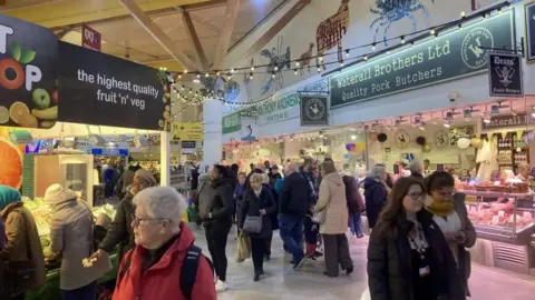 Victoria Scheer/BBC Crowds of people are shopping at food stalls at an indoor market.