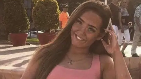 Anna Short A young woman with long dark hair and wearing a pink top sits smiling on a bench. She has her hand up next to her head. 
