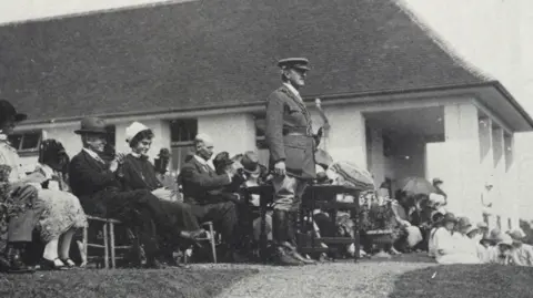 A picture of Field Marshal Viscount Allenby standing to address people gathered outside the hospital at its official opening, with seated dignitaries gathered behind him. 