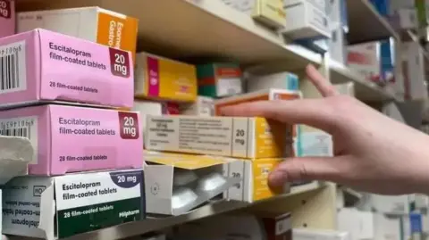 A shelf full of medication in colourful boxes, a hand goes to pick on box off the shelf.