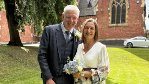 A couple stand in a church garden with the church behind them. The groom has white hair and is wearing a dark blue suit with a grey tie. The bride has short brown hair and a white and multi-coloured dress, she is carrying a blue bouquet. They are smiling at the camera. 