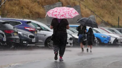 PA Media A woman walks with an umbrella. In front of her are two children also carrying an umbrella.