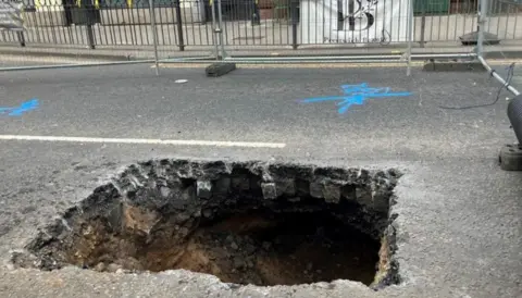 A hole appeared in Bury New Road. It is square-shaped and surrounded by fencing. 
