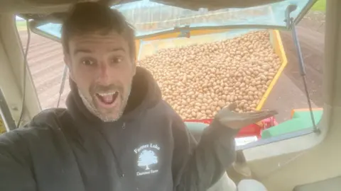 Luke Abblitt Luke, a man wearing a black hoodie, sitting inside a tractor pointing to his trailer full of potatoes. 