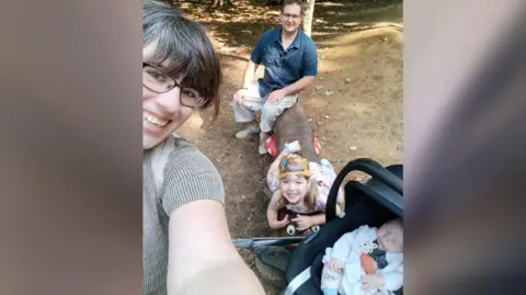 Adele Mann Selfie of Adele holding the phone - Oliver in a buggy, her daughter and husband sitting on a log