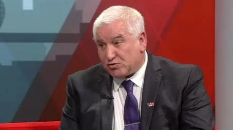 BBC A man with short white hair and wearing a dark suit, white shirt and purple tie, is sitting in a TV studio, looking towards the presenter out of shot to the left. He has a pin on the left lapel of his jacket with a black, white then red strip on it.