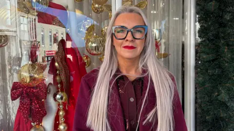 A woman with long blonde hair is smiling at the camera. She is wearing large dark rimmed glasses, red lipstick, and a burgundy top. Behind her is a Christmas display in the window of a shop.